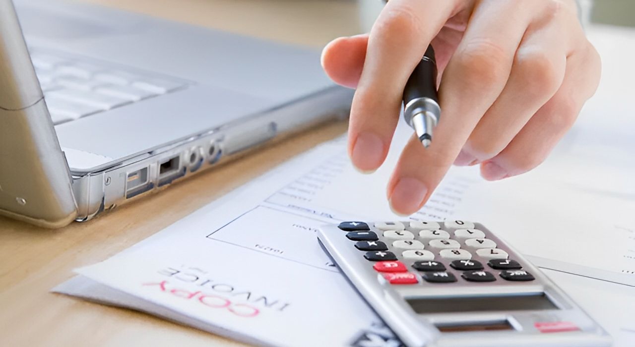 Person using a calculator and pen near a laptop on a desk.