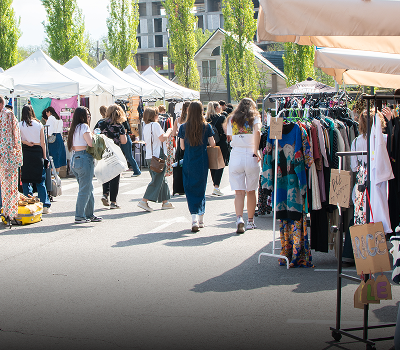 People browsing clothing racks and vendor booths at an outdoor market with white tents set up along a paved walkway on a sunny day.