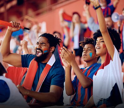 Three fans with painted faces cheer energetically in a stadium, wearing team colors and holding scarves, surrounded by other supportive spectators.