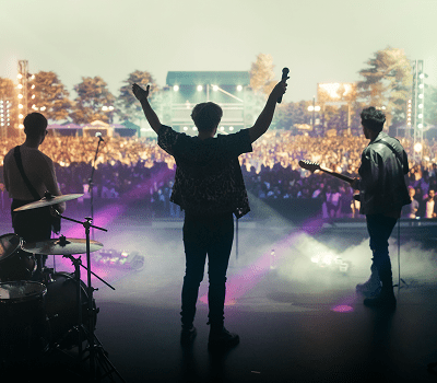 A band performs on an outdoor stage before a large, cheering crowd, with stage lights and musical instruments visible.