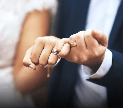 Close-up of two hands interlocking pinky fingers; one wears a diamond engagement ring, the other a wedding band. Both are dressed in formal wedding attire.