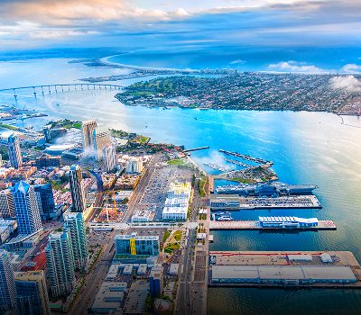 Aerial view of a coastal city with tall buildings, a curved bridge, and a harbor with piers extending into the blue water under a partly cloudy sky.