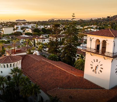 Aerial view of a Spanish-style building with a clock tower, red tile roofs, and surrounding trees at sunset in a coastal city.